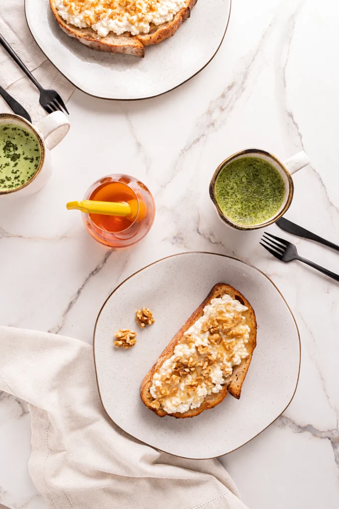 A imagem mostra um café da manhã visto de cima, servido sobre uma mesa clara com efeito marmorizado. Duas fatias de pão tostado aparecem cobertas por um creme branco, nozes e mel, acompanhadas por canecas com uma bebida verde. No centro, um recipiente de vidro com mel e um pegador amarelo completa a composição. As louças em tons neutros, os talheres escuros e o tecido cru criam uma cena leve, natural e acolhedora.