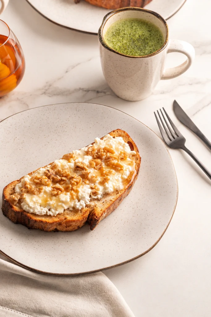 A imagem destaca uma fatia de pão tostado com cobertura cremosa, nozes e mel, servida em um prato bege de formato orgânico. Ao fundo, uma caneca com bebida verde, um recipiente com mel e talheres escuros completam a composição. A luz suave e os tons neutros criam uma cena de café da manhã leve e acolhedora.