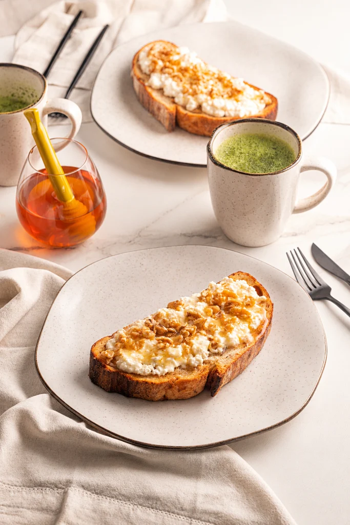 A imagem apresenta um café da manhã servido em louças de tom bege e acabamento pontilhado. Duas fatias de pão tostado levam uma cobertura cremosa, nozes e mel. Canecas com uma bebida verde, um recipiente de vidro com mel completa a composição. A iluminação suave, os tons neutros e o tecido claro criam uma cena leve, natural e acolhedora.