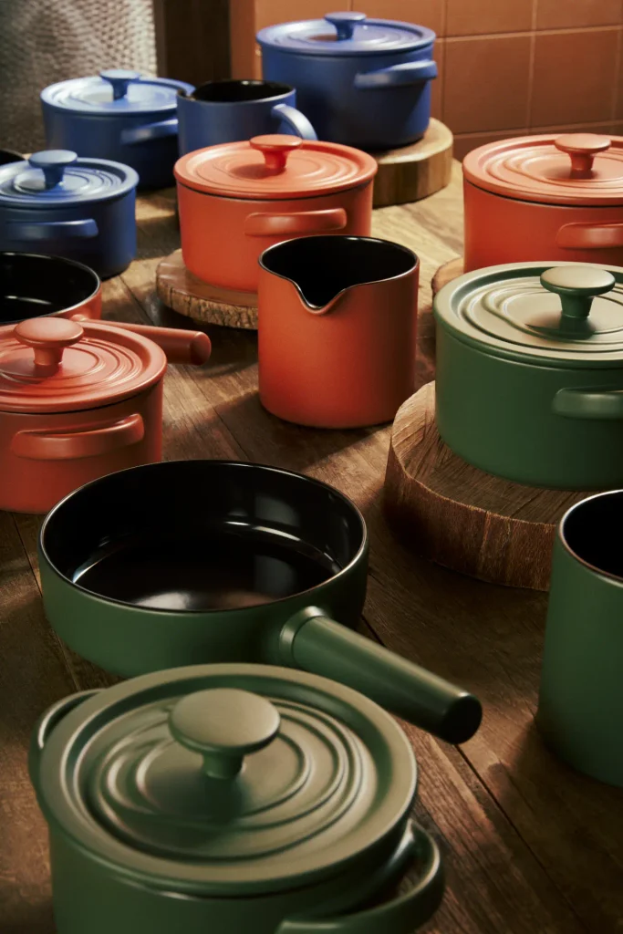 Panelas de cerâmica In Natura nas cores verde Taioba, coral Goiaba e azul Jenipapo distribuídas sobre uma mesa de madeira e bases feitas de fatias de tronco. A composição reúne caçarolas com tampa, fervedores com bico e uma frigideira verde com interior preto e cabo voltado para a direita. Ao fundo, aparecem azulejos em tons terrosos.