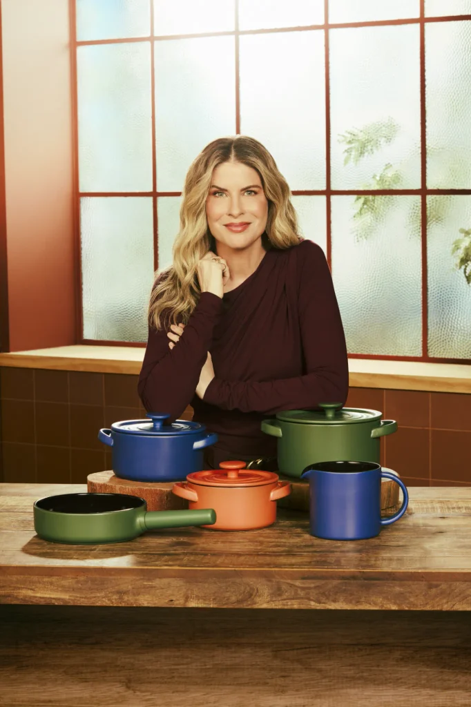 Rita Lobo com roupa vinho atrás de uma mesa de madeira, com uma mão apoiada sob o queixo. À frente, peças da coleção In Natura: frigideira verde Taioba, caçarola coral Goiaba com tampa, caçarolas azul Jenipapo e verde Taioba com tampa e fervedor azul com interior preto. A caçarola azul está elevada sobre uma base de madeira. Ao fundo, uma janela ampla com divisórias avermelhadas ilumina a cozinha.
