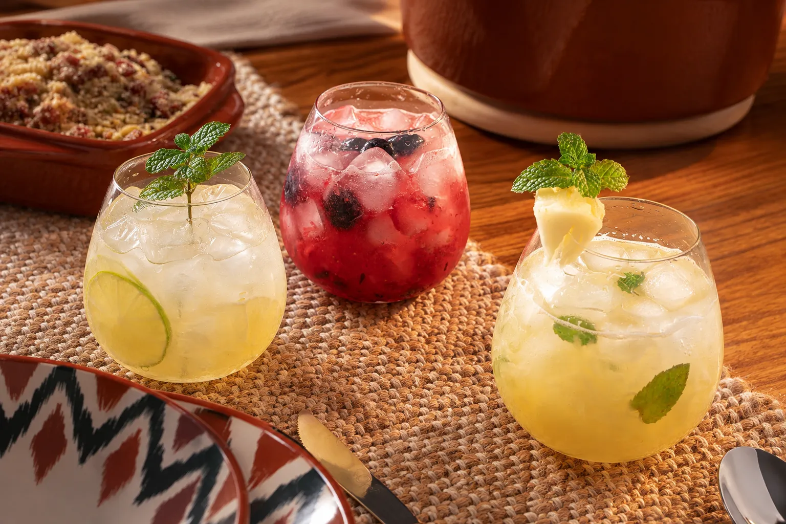 Três drinks sem álcool servidos em copos de cristal arredondados com gelo, sobre um caminho de mesa de fibra natural. À esquerda, bebida clara com rodela de limão e ramo de hortelã; ao centro, drink vermelho com pedaços de morango, amoras e mirtilos; à direita, bebida amarela de abacaxi com folhas de hortelã e um pedaço da fruta preso à borda do copo. Ao redor, há uma mesa de madeira, parte de um prato estampado, talheres e peças de cerâmica em tons terrosos.