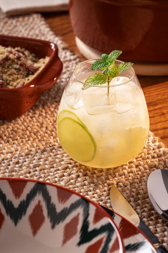Drink sem álcool de limão servido com bastante gelo em um copo baixo e arredondado, decorado com uma rodela de limão dentro da bebida e um ramo de hortelã na parte superior. O copo está sobre um caminho de mesa de fibra natural, acompanhado por parte de um prato estampado, talheres e peças de cerâmica em tons terrosos ao fundo.