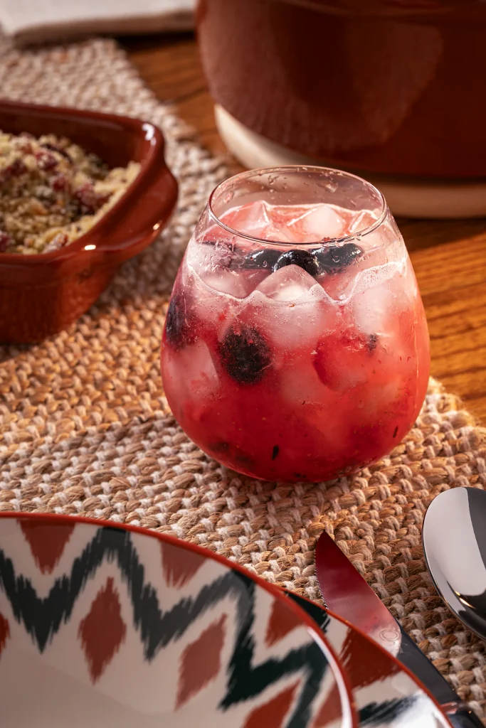 Drink sem álcool de frutas vermelhas servido em um copo baixo e arredondado, com bebida em tom rosa-avermelhado, bastante gelo, pedaços de morango, amoras e mirtilos. O copo está sobre um caminho de mesa trançado em tons naturais, em uma mesa de madeira. Ao redor, aparecem parte de um prato com estampa geométrica em verde, vermelho e bege, talheres metálicos e peças de cerâmica marrom, incluindo uma travessa com alimento e um recipiente maior ao fundo.