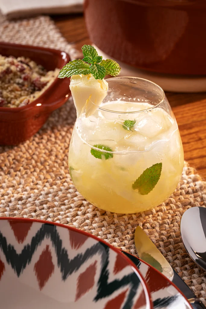 Drink de abacaxi em tom amarelo-claro, servido com bastante gelo em um copo baixo e arredondado. A bebida contém folhas de hortelã entre as pedras de gelo e está decorada com um pedaço de abacaxi e um ramo de hortelã na borda. O copo aparece sobre um caminho de mesa trançado em tons naturais, ao lado de talheres metálicos e de parte de um prato com estampa geométrica em verde, vermelho e bege. Ao fundo, peças de cerâmica marrom completam a composição sobre a mesa de madeira.
