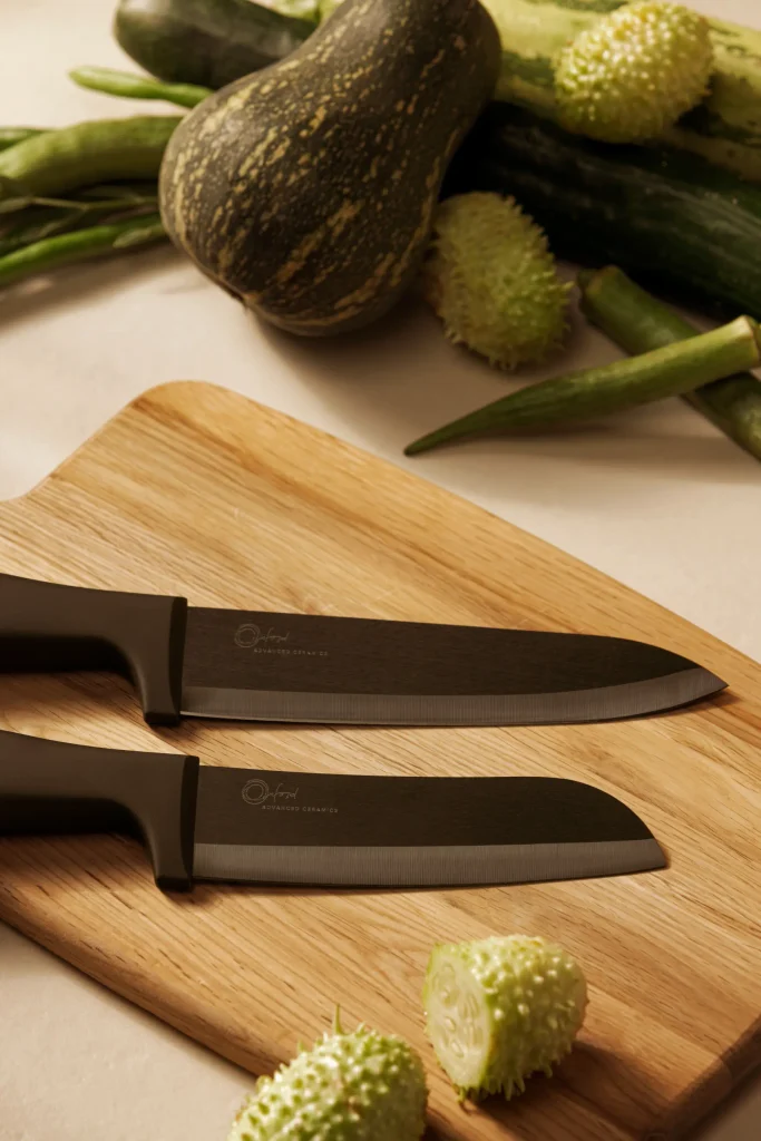 Vista aproximada das facas de cerâmica do Chef e Santoku Oxford All Black sobre uma tábua de madeira. As lâminas pretas apontam para a direita, com a faca do Chef acima e a Santoku abaixo. A marca Oxford aparece nas duas lâminas. Ao redor da tábua, há maxixes inteiros e cortados, abóbora, pepinos e quiabos sobre uma bancada clara, iluminada por luz natural.