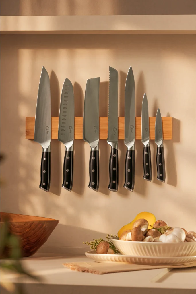 Sete facas Oxford com lâminas metálicas e cabos pretos com três rebites, alinhadas em um suporte de madeira na parede, com as pontas voltadas para cima. Entre os modelos, aparecem uma Santoku com cavidades ovais, um cutelo e uma faca de pão serrilhada. Na bancada abaixo, uma tigela clara sobre um prato reúne cogumelos, alho e pimentão amarelo, com ramos de ervas ao redor. Uma tigela de madeira aparece à esquerda. A luz quente projeta sombras das facas na parede bege.