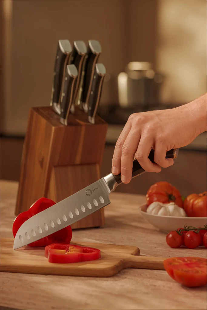 Mão segura uma faca Santoku Oxford de lâmina larga, com cavidades ovais, sobre um pimentão vermelho em uma tábua de madeira. Uma rodela de pimentão está à frente da lâmina. Ao fundo, um cepo de madeira acomoda facas com cabos escuros e detalhes metálicos. Tomates e uma cabeça de alho completam a composição sobre a bancada, iluminada em tons quentes.