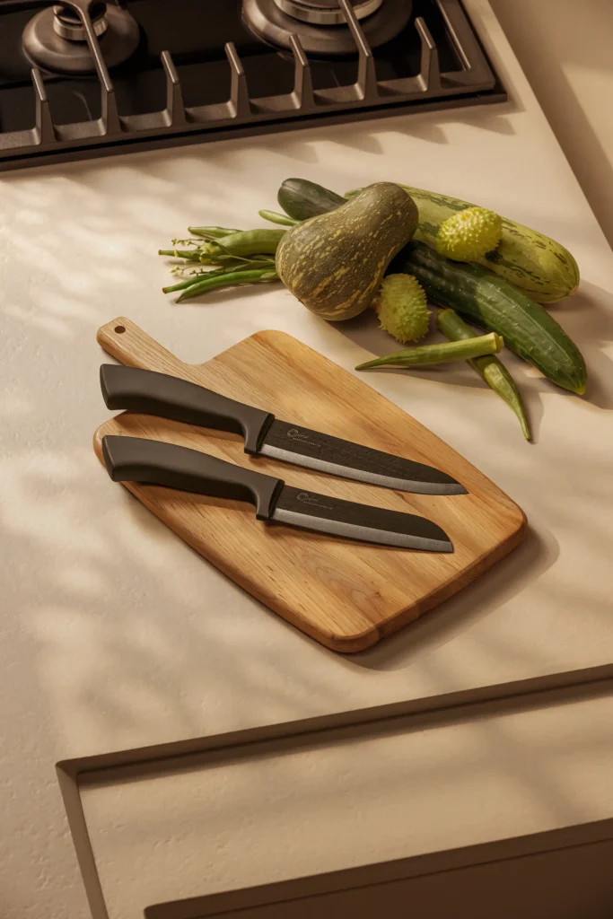 Facas de cerâmica do Chef e Santoku Oxford All Black lado a lado sobre uma tábua retangular de madeira com cabo. As facas têm cabos e lâminas pretos, com as pontas voltadas para a direita. A tábua está sobre uma bancada clara, próxima a um fogão a gás. Ao fundo, abóbora, pepinos, maxixes e quiabos formam uma composição iluminada pela luz natural, que projeta sombras suaves na bancada.