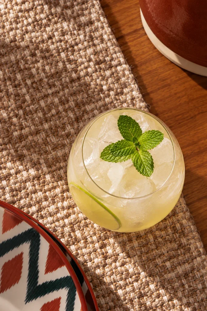 Vista de cima de um drink sem álcool de limão servido em um copo baixo e arredondado, com bastante gelo, uma rodela de limão dentro da bebida e um ramo de hortelã ao centro. O copo está sobre um caminho de mesa de fibra natural, em uma mesa de madeira, próximo a um prato com estampa geométrica colorida e uma peça de cerâmica marrom.